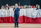 Chłopiec na scenie trzyma w ręce mikrofon i mówi. Z tyłu dzieci trzymają dużą biało czerwoną flagę.