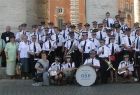 Orkiestra koncertuje w Rzymie. Muzycy w mundurach strażackich, w tle kolumny na placu Św. Piotra.