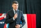 Wicemarszałek Łukasz Smółka trzyma w ręce mikrofon i przemawia. Z tyłu widać biało czerwoną flagę.