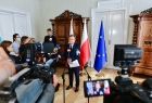 Marszałek podczas konferencji prasowej stoi na tle flag przed mikrofonami. Przed marszałkiem dzinnikarze