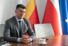 Wicemarszałek Łukasz Smółka siedzi przy stole, na tle trzech flag.