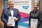 Przedstawiciel Małopolskiego Funduszu Rozwoju i Karolina Dera-Szymańska – Prezes Zarządu Małopolskiego Funduszu Rozwoju i prezes Funduszu Rozwoju i Promocji Województwa Wielkopolskiego trzymają w rękach teczki z umowami. Z tyłu widoczny baner Małopolska.