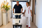 Na zdjęciu mężczyzna w średnim wieku korzystający z robota z funkcją chodzika rehabilitacyjnego. Obok młoda kobieta w białym fartuchu pielęgniarskim, która przechadza się wraz z mężczyzną korytarzem szpitala.