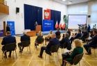 Goście uroczystości siedzą na krzesłach, widok z tyłu, przy mównicy stoi mężczyzna, za nim widoczne granatowe zasłony oraz godło RP