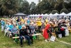 Seniorzy siedzą na ławkach w Parku Jordana. Z tyłu widoczna zieleń i białe namioty. 