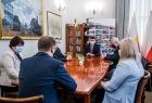 Grupa osób przy stole konferencyjnym podczas spotkania z wicemarszałkiem Łukaszem Smółką, w tle biblioteczka i flagi