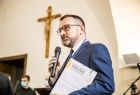 Marszałek Tomasz Urynowicz przemawia trzymając mikrofon i dyplom. W tle krzyż na ścianie.