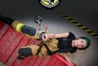 Grafika przedstawia kobietę w stroju strażackim, która wisi na linie. Zdjęcie wykonane jest z góry 