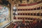 Zdjęcie widowni i kurtyny ozdobnej teatru. Na parterze ustawione są rzędy czerwonych krzeseł. Po lewej i prawej stronie umieszczone są złote, bardzo ozdobne balkony z miejscami dla widzów. Przed kurtyną stoi przewodnik teatralny. Na widowni siedzi grupa.