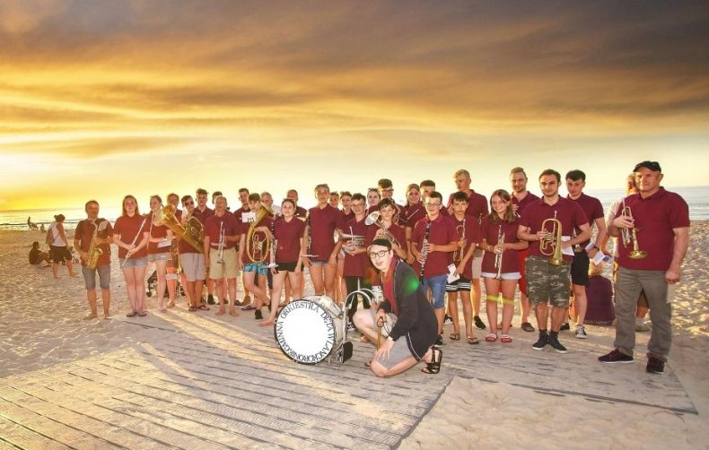 Gminna Orkiestra Dęta z Lanckorony stoi na plaży na tle zachodzącego słońca.