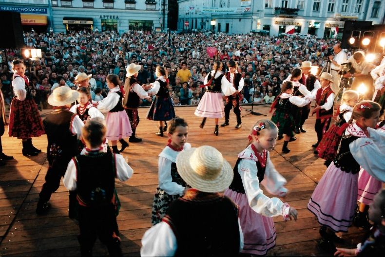 Kilka par dziecięcych w strojach regionalnych tańczących na scenie. W tle miejskie kamienice i widownia pod sceną