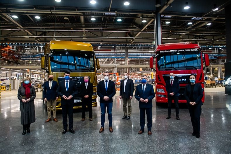 Marszałek Witold Kozłowski i wicemarszałek Łukasz Smółka stoją z innymi uczestnikami wydarzenia na hali produkcyjnej. Z tyłu widoczne dwie ciężarówki.