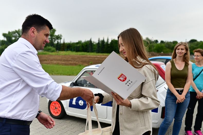 Wicemarszałek Łukasz Smółka wręcza dyplom młodej kobiecie. Z tyłu widoczny samochód.