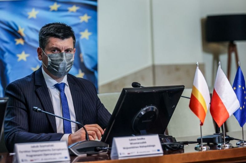 Wicemarszałek Łukasz Smółka siedzi na sali z maseczką na twarzy. Za nim baner Fundusze Europejskie.