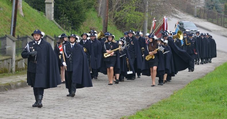 Orkiestra Dęta OSP Jordanów maszeruje ulicą w mundurach galowych.