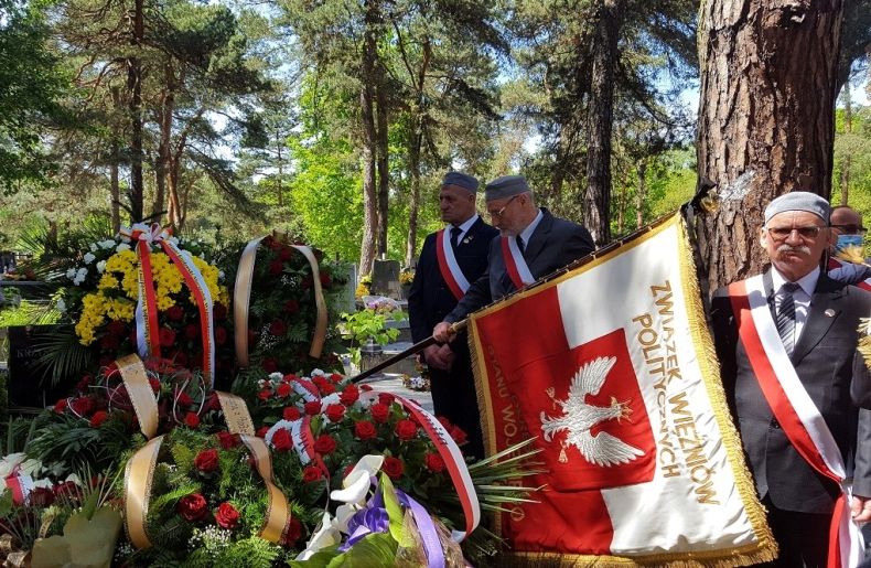 Uczestnicy uroczystości pogrzebowych przy grobie pokrytym kwiatami oraz wieńcami, na pierwszym planie mężczyzna trzymający sztandar Związku Więźniów Politycznych Okresu Stanu Wojennego