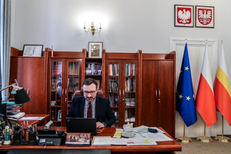 Wicemarszałek Tomasz Urynowicz przy biurku podczas posiedzenia Rady, w tle widoczne meble oraz trzy wysokie flagi stojące na podłodze po prawej stronie pomieszczenia