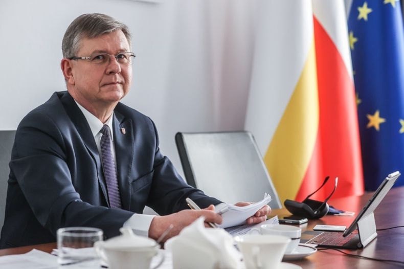 Marszałek Witold Kozłowski siedzi za biurkiem. Na stole urządzenie oraz dokumenty. W tle flagi: narodowa, wojewódzka i UE