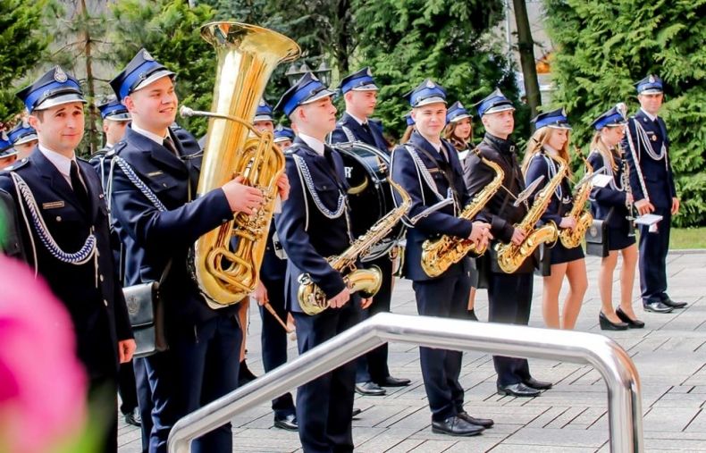 Orkiestra Dęta OSP Tokarnia stoi przed występem w mundurach strażackich. W tle zieleń.