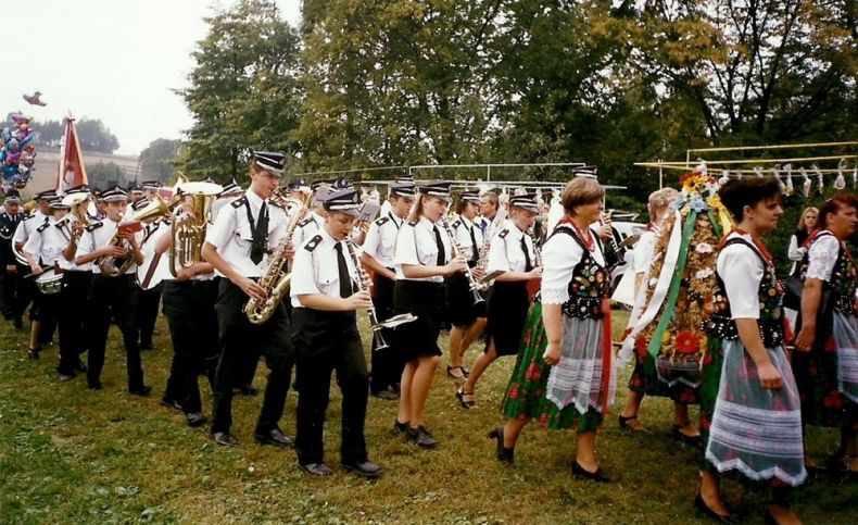 Orkiestra dęta podczas występu w plenerze. Maszeruje po trawie, przed nią kobiety w strojach ludowych.