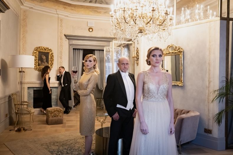 Na zdjęciu pięć osób w eleganckim salonie. Na Pierwszym planie kobieta w złotej sukni, obok mężczyzna w czarnym fraku i druga kobieta w złotej sukni. W tle przy kominku stoją dwie osoby. Kobieta w czarnej sukni i mężczyzna w czarnym garniturze.