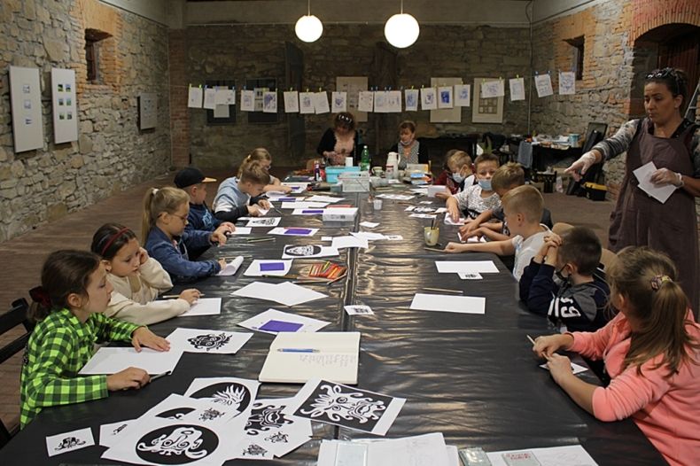 Na fotografii kolorowej są dzieci uczestniczące w warsztatach w ramach projektu pn. Szklany Renesans- dzieci wraz z nauczycielkami siedzą przy długim stole, instruktorka stoi obok