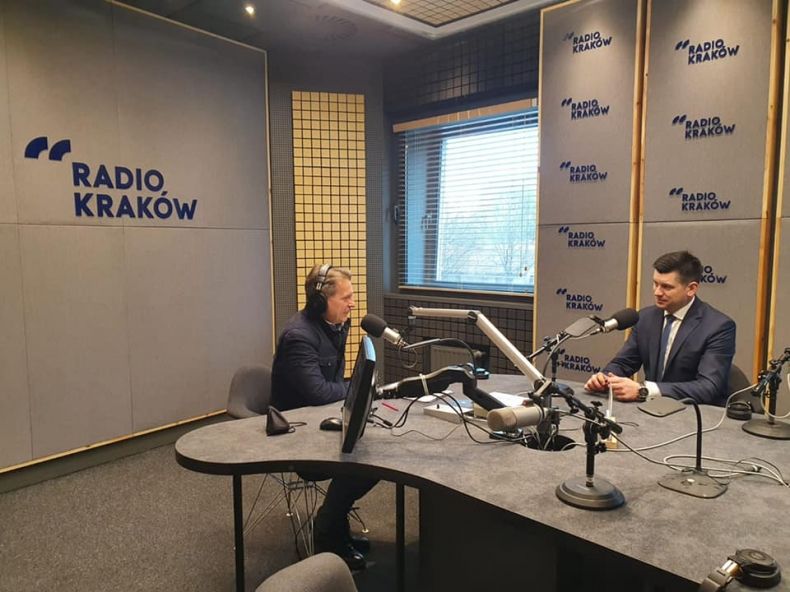 Wicemarszałek Łukasz Smółka siedzi przy stole z redaktorem w Radio Kraków. Z tyłu widoczna szara ściana z niebieskim logo Radia Kraków.