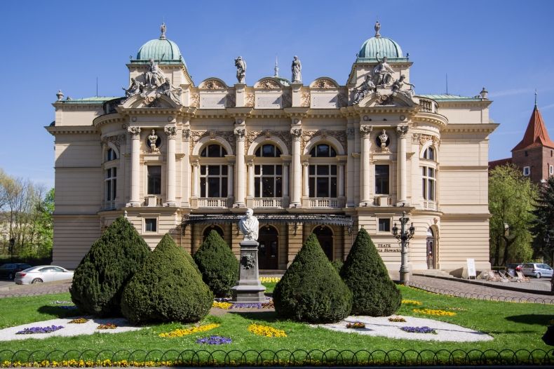 Na zdjęciu budynek Teatru imienia Juliusza Słowackiego w Krakowie. Fasada zdobiona rzeźbami. Duże okna. Przed wejściem zieleniec z krzewami w kształcie stożka. 