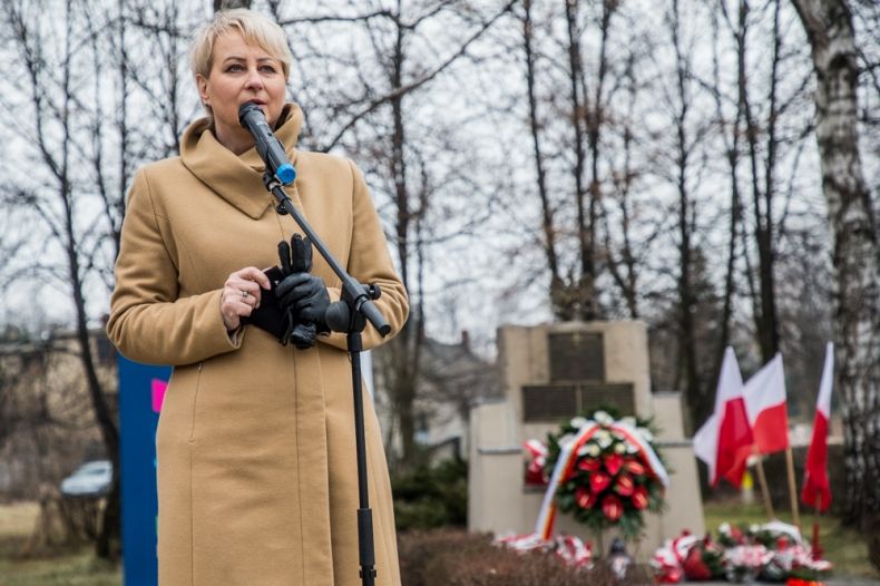 Iwona Gibas z zarządu województwa przemawia do mikrofonu. W tle widoczny pomnik oraz biało czerwone flagi.