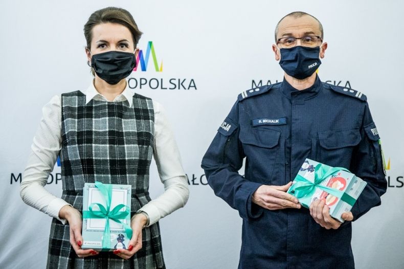 Marta Malec-Lech z zarządu województwa oraz Robert Michalik z Komendy Wojewódzkiej Policji w Krakowie stoją podczas konferencji i trzymają w dłoniach kartki świąteczne. W tle widoczny napis Małopolska.