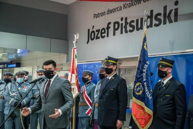 Wicemarszałek Łukasz Smółka przemawia podczas uroczystości na Dworcu Głównym w Krakowie. W tle napis Patron Dworca Kraków Główny Józef Piłsudski i poczty sztandarowe.