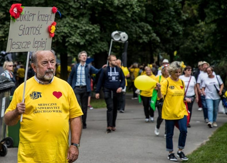 Seniorzy w czasie marszu Senior ma moc. Mężczyzna trzyma transparent z napisem Cukier krzepi starość jeszcze bardziej. Na żółtej koszulce ma napis Medyczny Uniwersytet Trzeciego Wieku.