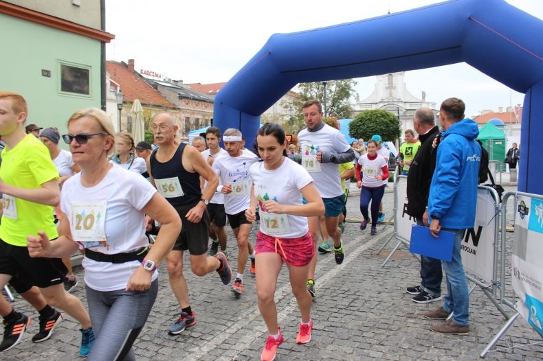 Zdjęcie przedstawia grupę biegnących osób, biorących udział w biegu ulicznym. W tle widoczna jest bramka startowa.