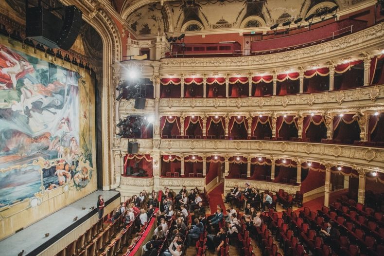 Zdjęcie widowni i kurtyny ozdobnej teatru. Na parterze ustawione są rzędy czerwonych krzeseł. Po lewej i prawej stronie umieszczone są złote, bardzo ozdobne balkony z miejscami dla widzów. Przed kurtyną stoi przewodnik teatralny. Na widowni siedzi grupa.