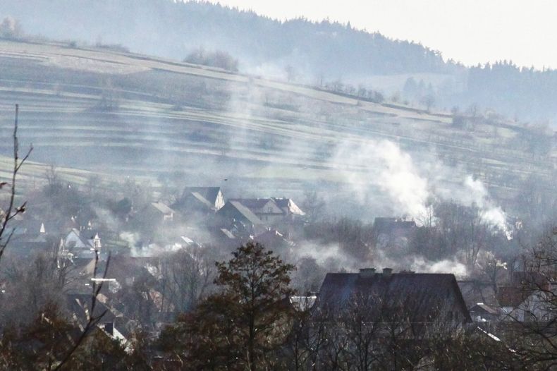 Widok na dachy domów spowite jasnoszarym dymem, w tle zielony krajobraz