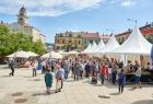 Widok na płytę gorlickiego rynku na którym ustawiono białe stożkowate namioty. W tle za nimi widać kolorowe kamieniczki, biały ratusz i zielone drzewa. Między namiotami widać spacerujących ludzi.