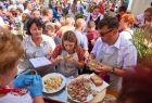 Radna Jadwiga Wójtowicz, stoi w grupie ludzi , przy stoisku. Za stoiskiem widać kobietę w stroju ludowym podającą stojącym w kolejce ludzią jedzenie. 