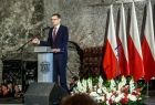 Premier RP Mateusz Morawicki stoi przy mównicy. Obok widoczne biało-czerwone flagi.