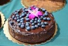 Widok na tort czekoladowy, ustawiony pośrodku zielonego obrusu. Jedno piętrowy tort, jest bogato oblany ciemnobrązową czekolada, posypany z wierzchu, borówka amerykańska i ozdobiony fioletowym kwiatem. 