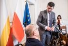 Wicemarszałek Łukasz Smółka rozmawia z niepełnosprawnym mężczyzną na wózku inwalidzkim. Obok widoczne flagi Małopolski, Polski i Unii Europejskiej.