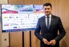 Wicemarszałek Łukasz Smółka stoi na tle telewizora z wyświetloną planszą IX Krakowskie Dni Bezpieczeństwa Ruchu Drogowego.