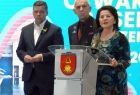Poseł Anna Paluch, poseł Jan Duda, senator Wiktor Durlak, wszyscy stoją przy szarym pulpicie, na którym umieszczono herb korzennej w kolorach czerwieni i złota. Za plecami mają niebieskie tło z częściowo widocznymi białymi napisami. 