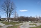 Widok na park w limanowej, w tle niebieskie niebo, z prawej strony widać wysokie drzewo. Trawnik poprzecinany jest szarymi ścieżkami. Na dalszym planie widać zabudowania i drzewa. 