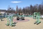 Park w limanowej, na pierwszym planie znajdują się zielone urządzenia do ćwiczeń, podłoże w kolorze żółtym w centralnym miejscu znajduję się okrąg w kolorze czerwonym, na nim okrągłe ławki i stolik. W tle widać niebieskie niebo i drzewa. 