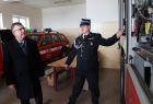 Wnętrze garażu OSP Wysokie. Marszałek Witold Kozłowski i prezes OSP Wysokie, stoją wewnątrz garażu. Strażak w galowym mundurze wskazuję na wyposażenie wozu strażackiego, które znajduję się po prawej stronie. W tle widać samochód osp w kolorze czerwony.