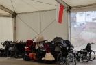 Wnętrze białego namiotu wypełnionego wózkami dla dzieci i sprzętem medycznym takim jak wózki inwalidzkie. Przy wejściu do namiotu widać czerwonobiała flagę. 