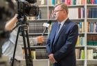 Marszałek Witold Kozłowski stoi na tle regałów wypełnionych książkami. Z lewej strony widać czarna kamerę ustawioną w kierunku marszałka, za nią sylwetkę dziennikarza w białej koszuli trzymającego w ręku mikrofon. 