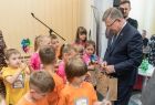 Marszałek Witold Kozłowski stoi w otoczeniu małych dzieci ubranych w pomarańczowe, żółte i różowe koszulki, marszałek wręcz dzieciom brązowe papierowe torby z upominkami. W tle za marszałkiem widać mężczyznę i kilka osób siedzących na krzesłach. 