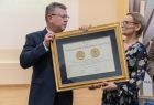 Marszałek Witold Kozłowski i dyrektor biblioteki Alina Marek. Marszałek Trzyma w ręku oprawioną w ramę w kolorze złota nagrodę Polonię Minor, którą przekazuję Alinie Marek. W tle widać żółte ściany pomieszczenia.