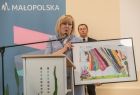 Radna Marta Mordarska, trzyma w ręku kolorowy obrazek, oprawiony w czarna ramkę. Radna stoi przy mównicy, tle widać rolap z napisem małopolska i kolorowym logo umieszczonym na niebieskim tle. 